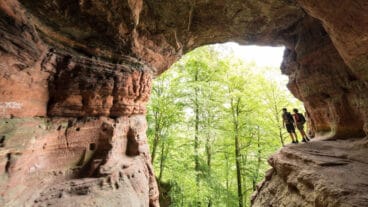 Two hikers at the mouth of the Genovevahöhle cave viewed from inside looking out at forest