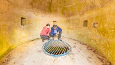 Two hikers kneeling in a round, moss-covered room, looking down at the Lieser spring under a blue grate